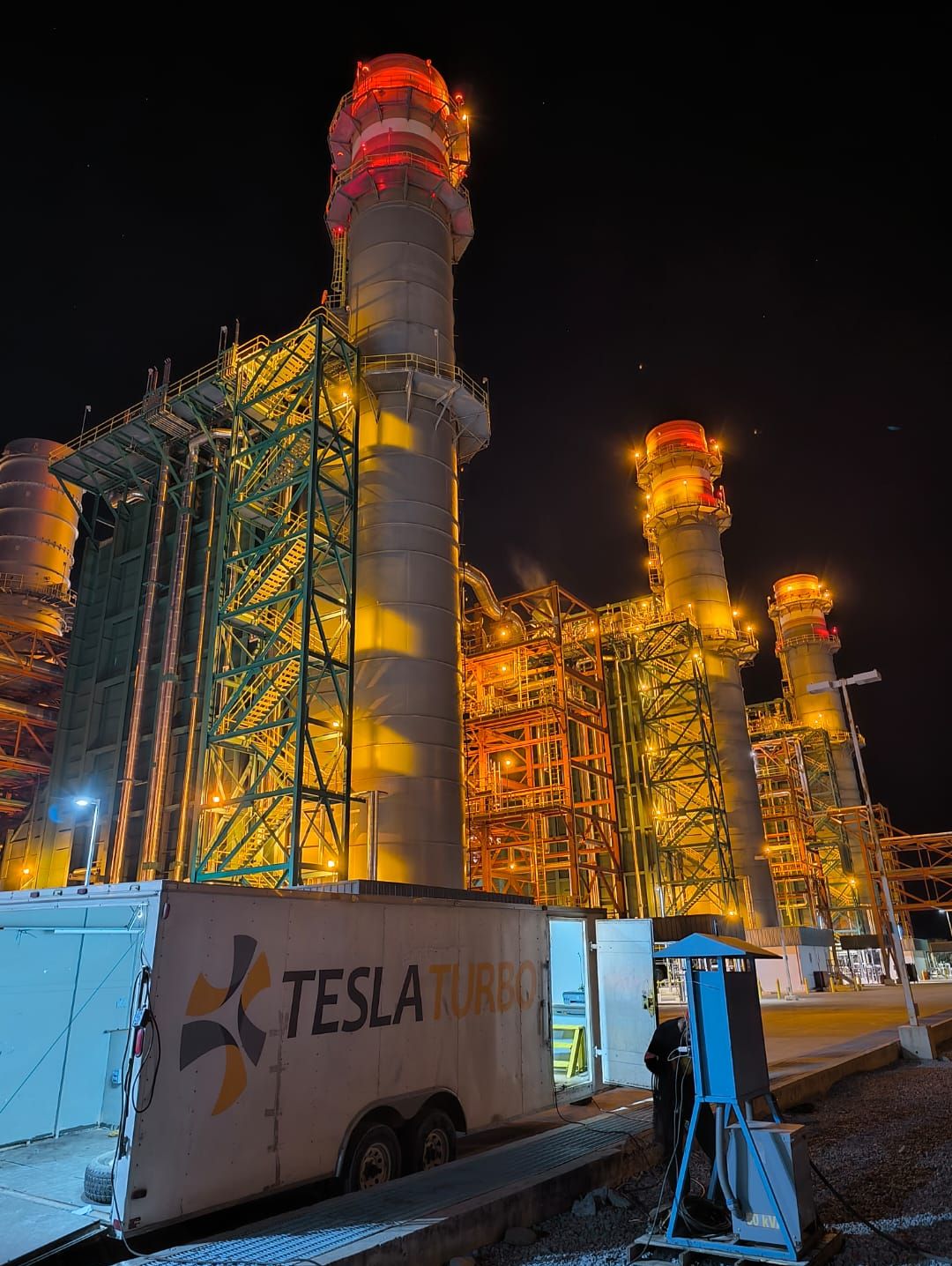 Unidad de Tesla Turbo junto a las chimeneas de una central de ciclo combinado, de noche
